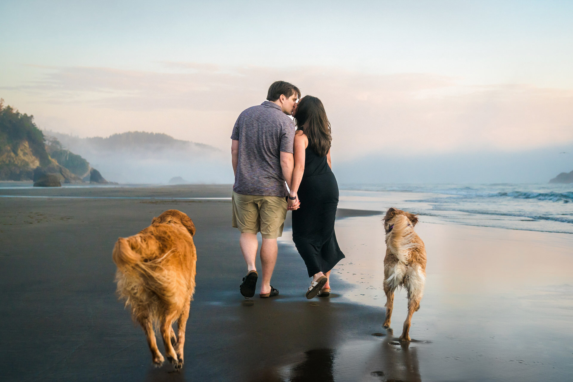 Couple kisses while their dogs follow behind at the beach in Oregon