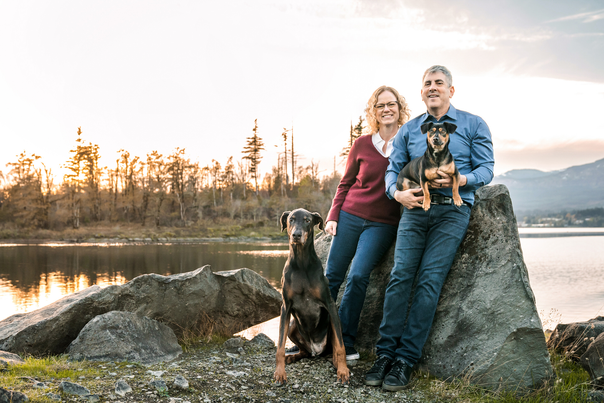Couple stands with their dogs by the water at sunset